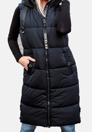 Kvinde iført en sort, ærmeløs polstret vest med en frontlynlås, hætte og hænder i lommerne over en sort langærmet skjorte og jeans.
