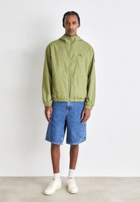 Giacca leggera verde chiaro con zip e cappuccio, abbinata a pantaloni corti di jeans blu e sneakers bianche. Design semplice e vestibilità casual.