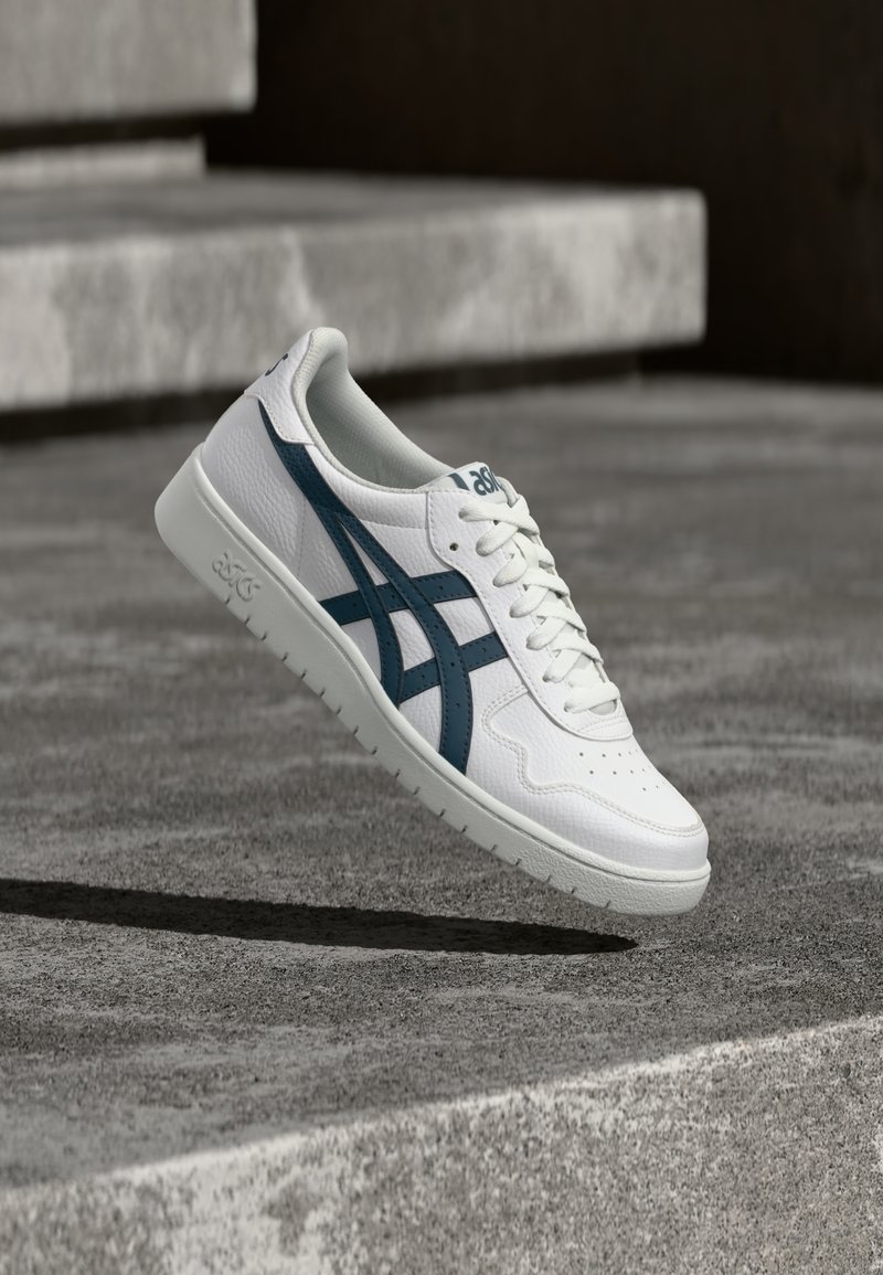 Valge sportlik king, millel on heledashall tald, sinised triibud ja perforatsioonid hingavuse tagamiseks. Sile tekstuur ja paelaga disain.