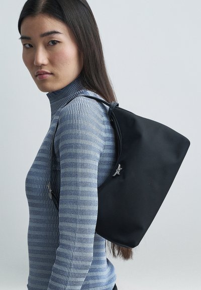 Jauna sieviete, ģērbusies zilā svītrā turtlenecka džemperī, nēsā lielu melnu plecu somu ar sudraba tauriņa rotājumu.