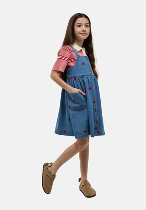 Jeune fille aux longs cheveux bruns portant un t-shirt rayé rose sous une robe en jean boutonnée avec des motifs de cœurs rouges et des chaussures marron sans lacets.