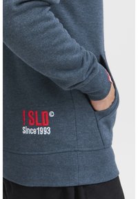 Persona che indossa una felpa blu con una mano in tasca e "!SLD Dal 1993" ricamato in rosso e bianco vicino alla vita.