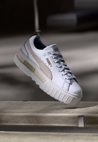 Sneaker bianco Puma con striscia in suede beige, suola spessa, loghi Puma dorati, lacci legati, posizionata in aria sopra una superficie di pietra.