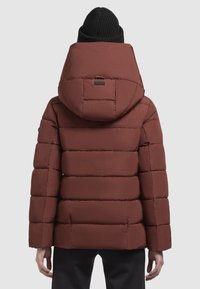 Braune gepolsterte Jacke mit einer großen Kapuze, gestepptem Muster und Seitentaschen. Getragen mit einer schwarzen Strickmütze.