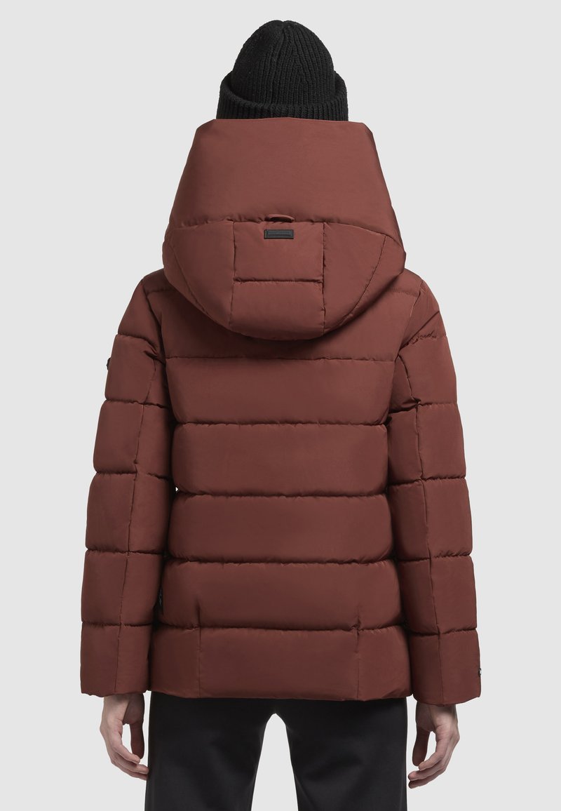 Veste marron rembourrée avec une grande capuche, texture matelassée et poches latérales. Portée avec un bonnet en tricot noir.