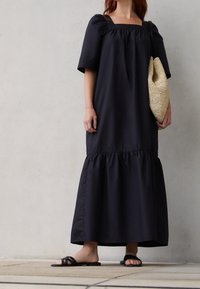 Robe maxi noire texturée avec des manches bouffantes, décolleté carré et ourlet volanté. Stylisée avec un sac tissé beige et des sandales plates noires.