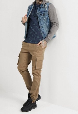 Hombre con pantalones cargo color beige, zapatillas negras, camisa azul marino con estampado, sudadera gris y chaqueta vaquera sin mangas, de pie contra una pared blanca.