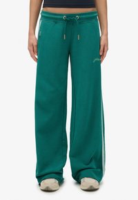 Persoon in groene, wijde sweats met een tailleband met trekkoord en sneakers, staand tegen een witte achtergrond.