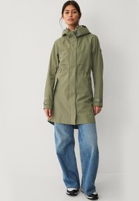 Giacca impermeabile verde oliva con cappuccio, vita stretta e due tasche frontali. Abbinata a jeans blu larghi e sneakers nere.