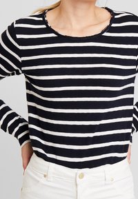 Maglietta a maniche lunghe a righe blu navy e bianche con scollo rotondo e orlo sfrangiato. Realizzata in un materiale morbido e leggero, abbinata a pantaloni bianchi.