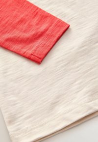 Helles T-Shirt mit weicher Textur; hat einen korallenroten Ärmel. Verfügt über einen runden Halsausschnitt und eine Naht entlang dem Saum.