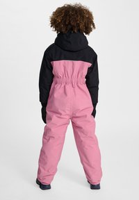 Roze en zwarte skisuit met een getailleerde taille, capuchon en lange mouwen. Beschikt over een rugpand en elastische manchetten. Gemaakt van een gestructureerde, duurzame stof.
