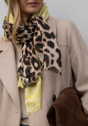 Frau trägt einen beigen Mantel mit geschichteten gelben und Leopardenmuster-Schals und hält eine braune Wildledertasche unter dem Arm.