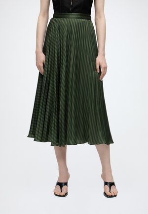 Femme portant une jupe midi plissée vert olive avec un top noir et des sandales à talons ouvertes noires, debout sur fond blanc.