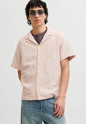 Ung mand iført grå solbriller, en kortærmet lyserød mønstret skjorte med knapper over en mørk T-shirt og blå jeans, stående mod en hvid baggrund.