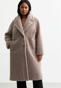 Cappotto in pelliccia sintetica beige con colletto a scialle, maniche lunghe e tasche laterali. Presenta una superficie testurizzata e una chiusura con un bottone.