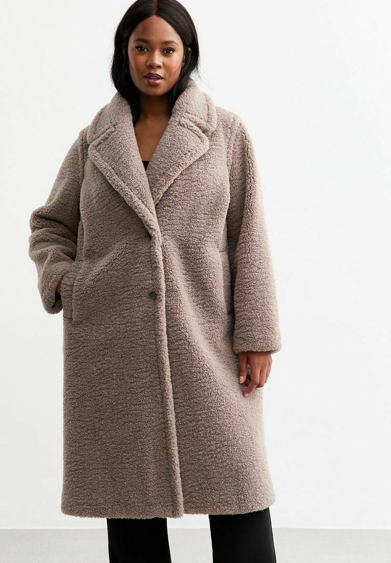 Cappotto in pelliccia sintetica beige con colletto a scialle, maniche lunghe e tasche laterali. Presenta una superficie testurizzata e una chiusura con un bottone.