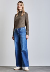 Vihreä oliivinvärinen pitkähihainen paita yhdistettynä leveisiin sinisiin farkkuihin ja valkoisiin tennareihin. Farkuissa on klassinen viisi-taskuinen muotoilu ja sininen pesu.