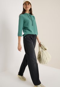 Top verde texturizado con capucha, pantalones de pierna ancha negros y zapatillas beige. Llevando un bolso suave de color claro.
