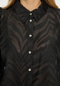 Melnas caurspīdīgas blūzes ar zickzaga rakstu, apkaklīti un pogu aizdari ar spīdīgām pogām. Redzams teksturēts auduma detaļas.