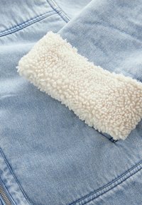 Tessuto denim blu chiaro con una morbida patch in pile crema. Il pile ha una superficie testurizzata e sollevata ed è cucito sul denim.