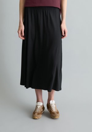 Femme portant une jupe midi noire associée à des baskets marron et beige, debout devant un fond uni gris clair.