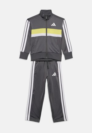 Tmavosivá tepláková súprava Adidas so zapínateľnou bundou a nohavicami s bielymi a žltými pásmi a bielymi logami Adidas na bunde a nohaviciach.