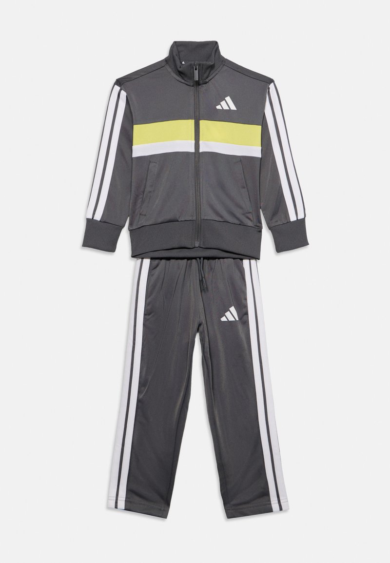 Σκούρο γκρι φόρμα Adidas με μπουφάν με φερμουάρ και παντελόνι με λευκές και κίτρινες ρίγες και λευκά λογότυπα Adidas στο μπουφάν και στο παντελόνι.
