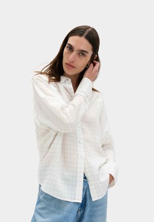 Kvinde med langt mørkt hår iført en hvid ternet skjorte med knapper og blå jeans, der rører ved sit hår med den ene hånd i en neutral setting.