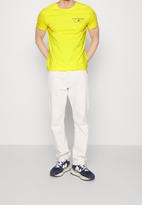 Muž v žlutém tričku Tommy Hilfiger, krémových kalhotách a modro-bílých teniskách stojí se založenýma rukama za zády.