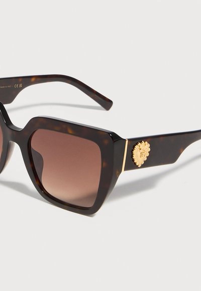 Τετράγωνα γυαλιά ηλίου με φίλτρα καφέ βαθμωτής απόχρωσης και χρυσό έμβλημα στο μπράτσο, τοποθετημένα σε λευκή επιφάνεια.