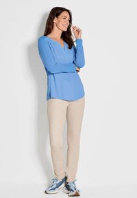 Langärmliges hellblaues Oberteil mit V-Ausschnitt, kombiniert mit beigen Hosen und blauen Schuhen, vor einem weißen Hintergrund.