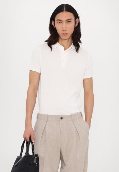 Polo shirt blanc à manches courtes avec un col à deux boutons, associé à un pantalon beige clair plissé. Le mannequin tient un sac à main noir.
