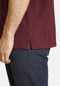 Burgundy cotton t-shirt with side slits, visible stitching, and short sleeves, paired with dark blue jeans. Close-up of fit and texture.