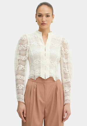 Donna che indossa una camicetta bianca in pizzo con maniche lunghe e orlo smerlato e pantaloni beige a pieghe a vita alta, in piedi davanti a uno sfondo semplice.