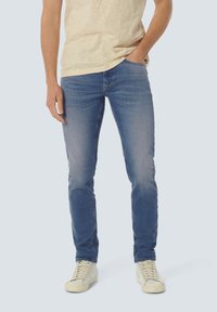 Strakke blauwe jeans met een vervaagde afwerking, vijf-pocket ontwerp en een klassieke tailleband, gecombineerd met witte sneakers en een lichtgekleurde shirt.