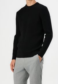 Suéter negro de punto acanalado con cuello redondo, combinado con pantalones a rayas en gris claro, que presentan una silueta ajustada y una textura sutil.