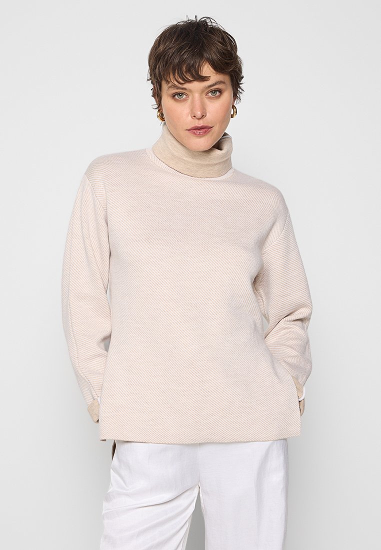 Beige Rollkragenpullover mit strukturiertem, geripptem Design. Lässiger Schnitt, lange Ärmel und seitliche Schlitze. Getragen mit weißen Hosen.