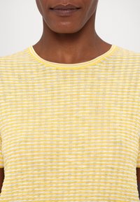 Detail osoby nosící žluté a bílé tričko s vodorovnými pruhy a krátkými rukávy s kulatým výstřihem.