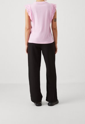 Kobieta ubrana w jasnoróżowy bezrękawnik z falbanami z dziurkowanym wzorem na rękawach, czarne szerokie spodnie i czarne buty, widziana od tyłu.