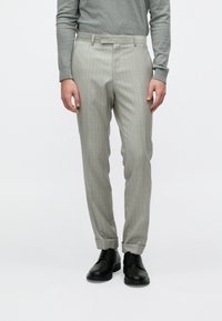 Mand iført lysegrå bukser med nålestriber, grå langærmet skjorte og sorte snørebånds-sko stående mod en ensfarvet baggrund.