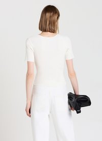 Top fitted bianco a maniche corte con scollo rotondo, abbinato a jeans bianchi a vita alta. La modella tiene una borsa nera.