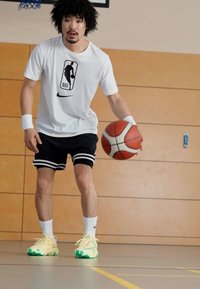 Basketbalista dribluje míč, má na sobě bílé tričko s logem NBA, černé šortky se pruhy a jasně zeleno-žluté tenisky.