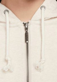 Ljusbeige hoodie med en silverfärgad dragkedja med en "NIKE"-tagg, vita dragsnören med tofsar och ett mjukt texturerat tyg.