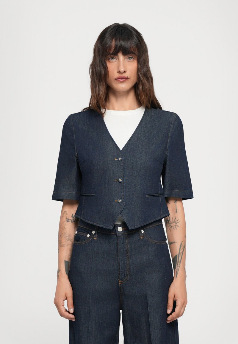 Gilet in denim corto con maniche corte, colore blu scuro, scollatura a V e chiusura frontale con cinque bottoni. Presenta due tasche laterali e cuciture a contrasto.