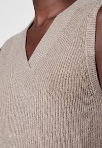 Pull sans manches beige en maille avec encolure en V et texture côtelée, présentant une coupe ajustée et des détails lisses.