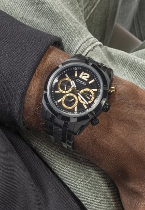 Orologio da polso GUESS nero e oro con tre quadranti secondari e cinturino in metallo, indossato sul polso sinistro sopra abbigliamento casual.
