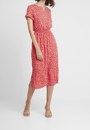 Mujer con vestido midi color coral con estampado de puntos blancos, mangas cortas, cintura fruncida y sandalias beige de tacón y punta abierta, de pie sobre fondo blanco.