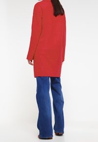 Rode lange jas met een rechte snit en strakke lijnen, gecombineerd met blauwe flared jeans. De stof lijkt zacht en glad.