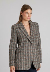 Blazer de cuadros grises con un corte entallado, que presenta un escote en V profundo, hombros estructurados y una tela tejida texturizada, acentuada con hilos marrones y blancos.